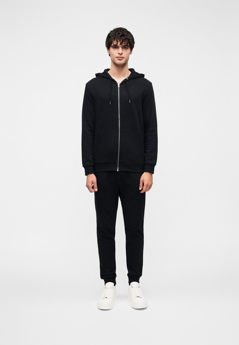 Μαύρο φούτερ με φερμουάρ, κουκούλα με κορδόνι και μπροστινές τσέπες, σε συνδυασμό με μαύρη φόρμα που ταιριάζει και λευκά αθλητικά παπούτσια. Ομαλή υφή.