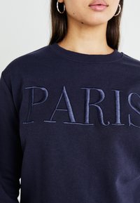 Sweatshirt azul-marinho com a inscrição "PARIS" em letras bordadas. Tem um decote redondo e uma textura suave. O tecido parece liso e resistente.