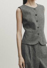 Harmaa villaliivi, jossa on sulava siluetti, pyöreä pääntie ja viisi etunappia. Ilman hihoja; sisältää hienovaraisen tekstuurin ja kaksi sivutaskua.