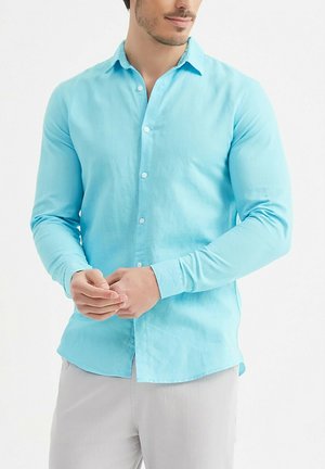 Homme portant une chemise à manches longues bleu clair boutonnée et un pantalon gris clair, les mains jointes devant lui.