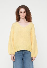 Γυναίκα με σγουρά κόκκινα μαλλιά, φοράει χαλαρό κίτρινο πλεκτό πουλόβερ και μπλε τζιν, στέκεται μπροστά σε λευκό απλό φόντο.