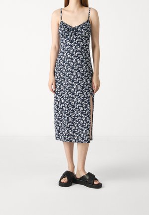 Kvinde iført en navyblå midi-kjole med hvidt blomsterprint, tynde stropper, sideslids og sorte platform-sandaler, stående foran en hvid baggrund.