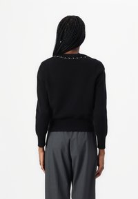 Czarny sweter, ściągacz na dole, dekolt okrągły, ozdobiony dekoracyjnymi ćwiekami wzdłuż linii szyi, noszony z szarymi spodniami, tkanina o fakturze.