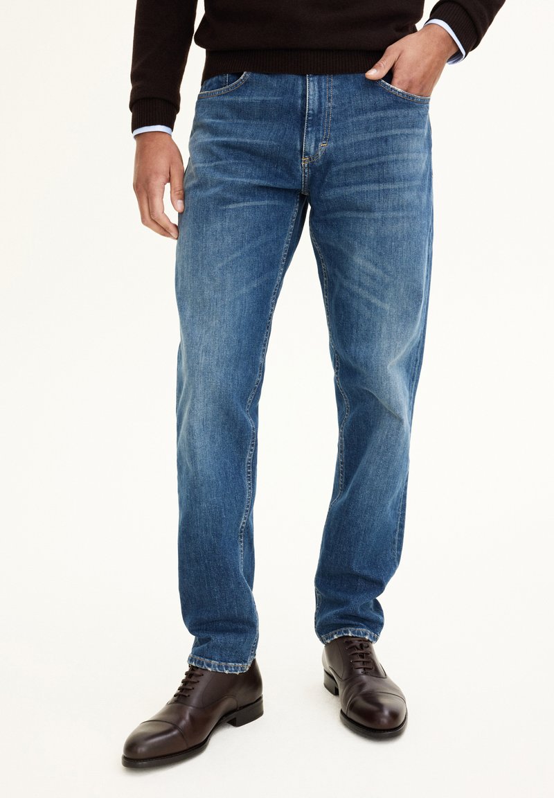 Blaue Denim-Jeans mit geradem Schnitt, ausgestattet mit einem Fünf-Taschen-Design und subtilen Used-Effekten. Kombiniert mit dunkelbraunen Lederschuhen.