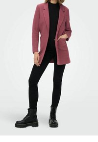 Veste rose en tissu texturé, design à simple boutonnage, deux poches avant ; associée à un pantalon noir ajusté et des bottines noires à lacets.