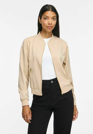 Femme aux longs cheveux foncés portant une veste beige zippée sur une chemise blanche et un pantalon noir, debout devant un fond blanc uni.