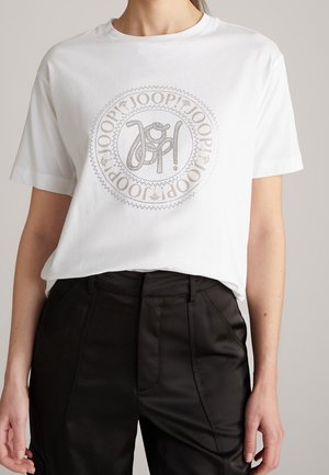 Femme portant une chemise blanche à manches courtes avec un logo circulaire "JOOP!" en strass, associée à un pantalon noir taille haute.