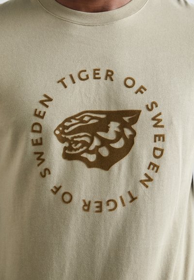 Olīvzaļa t-krekls ar teksturētu brūnu tīģera grafiku un aplī esošo "TIGER OF SWEDEN" tekstu, ar apaļu kakla izgriezumu un īsām piedurknēm.