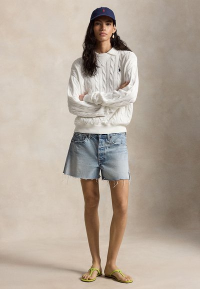 Ženska, oblečena v belo pulover s pletenim vzorcem, svetlo denim kratke hlače, mornarsko modro kapo in limetasto zelene japonke, stoji z rokami prekrižanimi pred enobarvnim ozadjem.