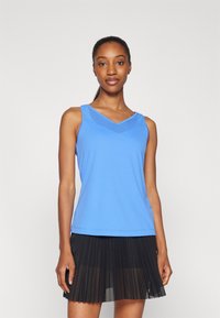 Donna che indossa un top senza maniche blu con dettagli in rete e una gonna plissettata nera, in piedi contro uno sfondo bianco semplice.