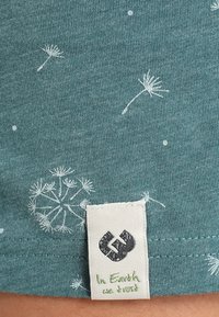 Teal fabric with white dandelion seed prints and a white tag showing a cracked heart icon and green text saying "In Earth we trust."