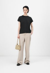 Svart t-shirt med kort ärm, matchad med beige byxor med vid ben. Modellen håller en liten, ljus beige handväska. Svarta skor med teksturerad design.