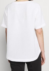 Vit kortärmad topp med en strukturerad, något voluminös tyg och rundad nederkant. Har en lös passform och enkel design.