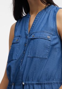 Světle modrá denimová bezrukávová blůza s výstřihem do V, předním zapínáním na knoflíky a dvěma náprsními kapsami se zmáčknutím.