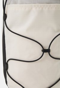 Bežový batoh z látky s černým elastickým šňůrkovým provázkem zajištěným černým plastovým uzávěrem na přední kapse.