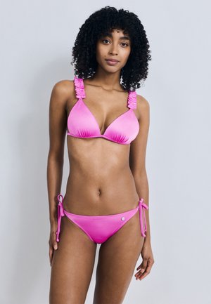 Γυναίκα με σγουρά μαλλιά που φοράει φωτεινό ροζ μπικίνι με βολάν στους ιμάντες, στέκεται μπροστά σε απλό ανοιχτό γκρι φόντο.