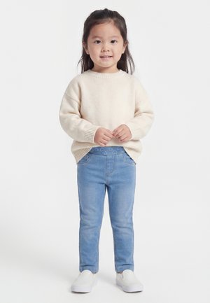 Jeune enfant debout, portant un pull crème, un jean bleu clair avec une ceinture à motif de cœurs et des chaussures blanches à enfiler sur un fond neutre.