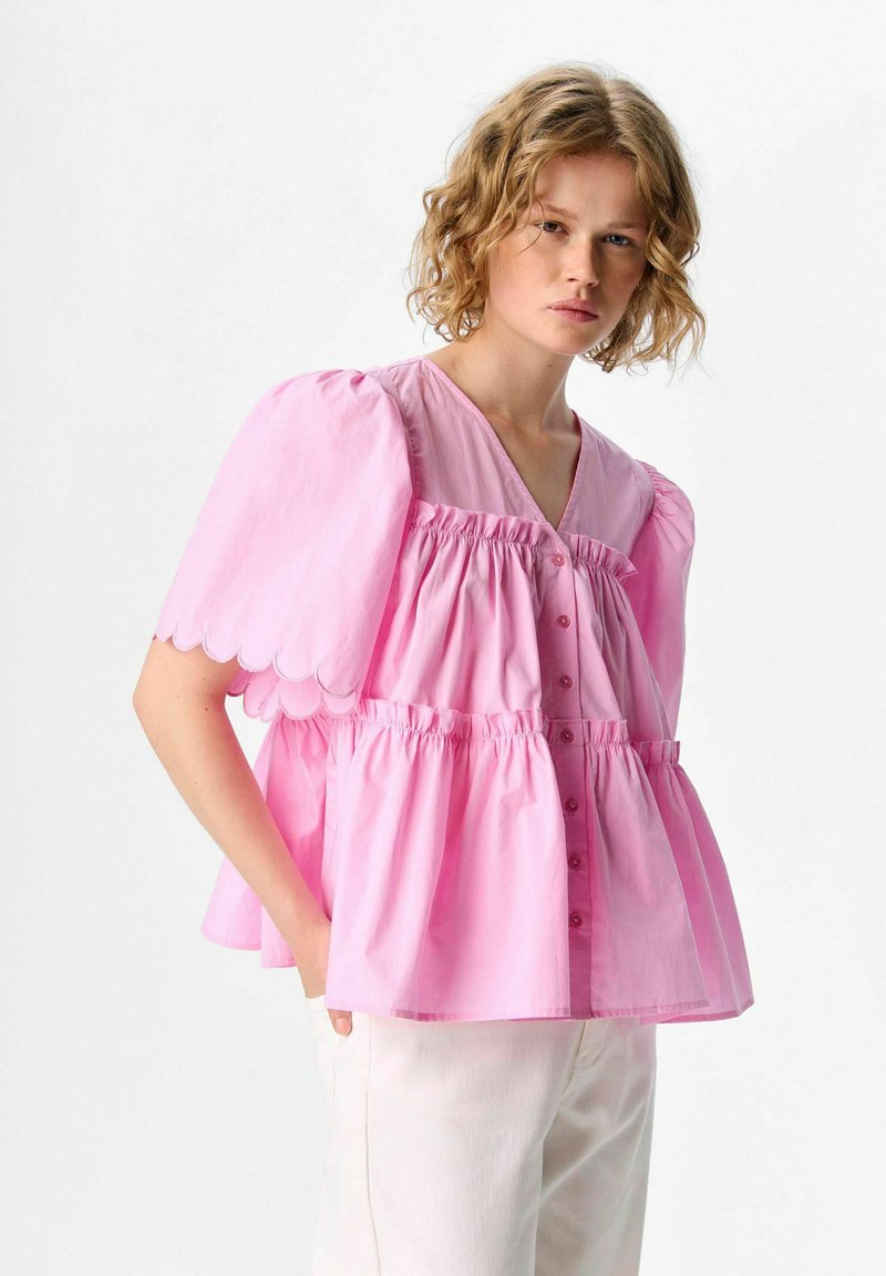 Blusa rosa a maniche corte con scollatura a V, strati arricciati e bordi delle maniche scallopati. Presenta una chiusura con bottoni e un tessuto leggero.