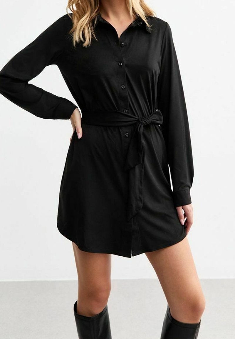 Abito camicia nero con maniche lunghe, apertura frontale con bottoni, cintura legata in vita e texture del tessuto liscia. Taglio lungo fino al ginocchio.