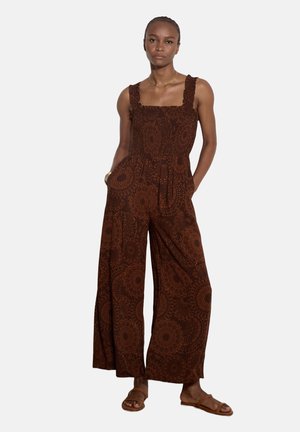 Donna che indossa una tuta marrone senza maniche con gambe ampie e motivo a cerchi, abbinata a sandali marroni bassi.