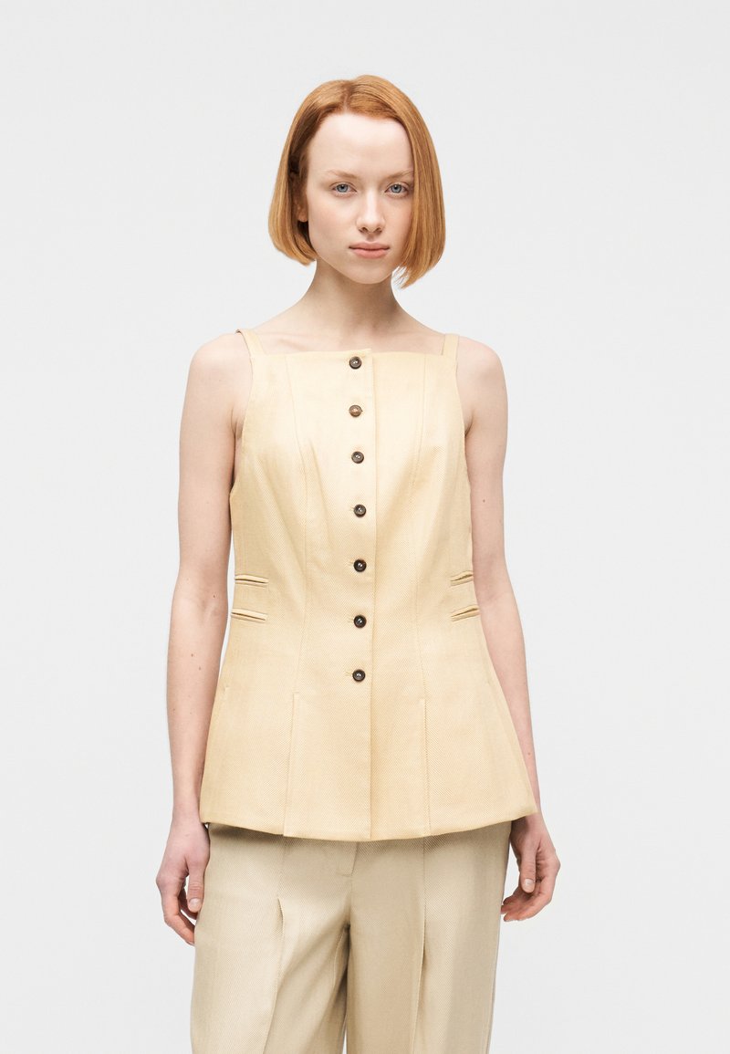 Νεαρή γυναίκα με κοντά κόκκινα μαλ λιά φοράει αμάνικο μπεζ τοπ με κουμπιά μπροστά και ταιριαστό μπεζ παντελόνι, στέκεται σε λευκό απλό φόντο.