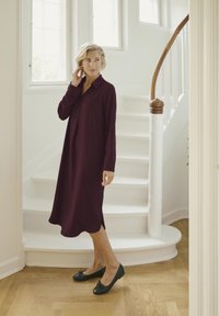 Robe bordeaux à manches longues avec un col boutonné, des fentes latérales et une coupe décontractée. Portée avec des ballerines noires. Escalier en colimaçon blanc en arrière-plan.