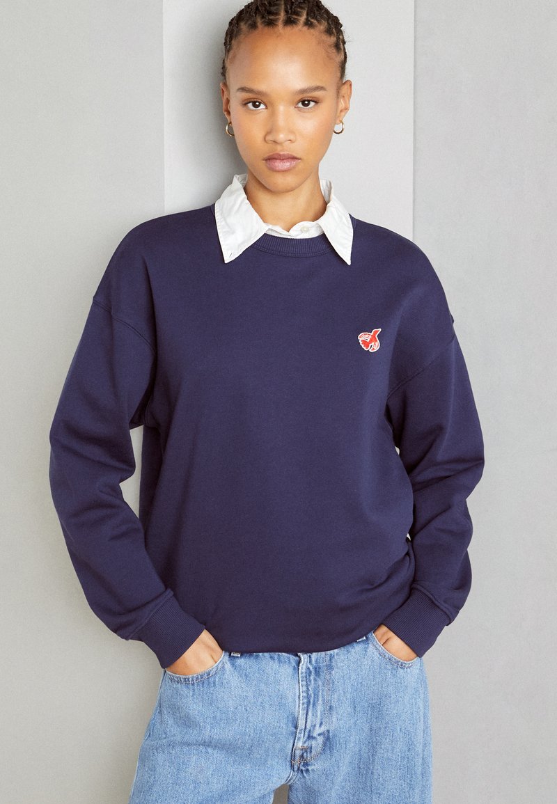 Σκούρο μπλε φούτερ με λαιμόκοψη, μακριά μανίκια και ένα μικρό κόκκινο λογότυπο στο στήθος, φορεμένο πάνω από ένα άσπρο πουκάμισο με γιακά.