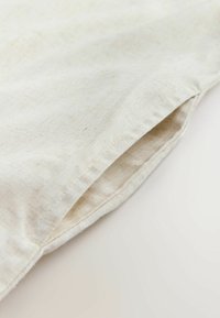 Tissu en lin naturel de couleur crème, présentant une texture subtile et un bord partiellement plié. Il comporte un ourlet brut et une apparence douce et aérée.