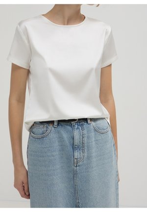 Femme portant un chemisier blanc uni à manches courtes rentré dans une jupe en denim bleu clair taille haute avec une ceinture noire.