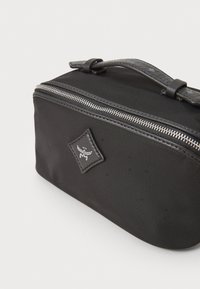 Sac en tissu noir avec une forme arrondie, fermeture éclair argentée, patch logo en relief et une poignée texturée. Matériau lisse avec un léger éclat.