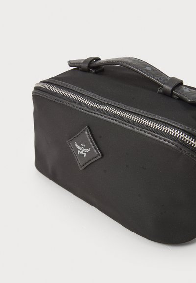 Mustast nailonist kosmeetikakotist, millel on ümar kuju, hõbedane lukku, tekstuuriga käepide ja väike logopatch.