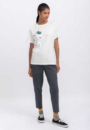 T-shirt bianca in cotone con una grafica di un'auto blu e accenti colorati sparsi, abbinata a pantaloni grigi e sneaker nere.