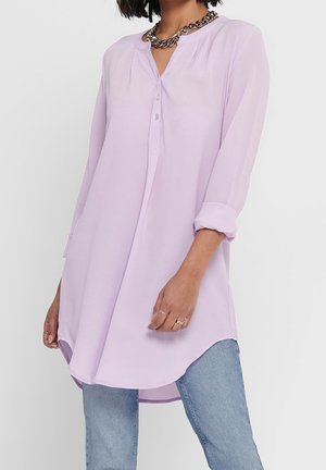 Femme portant une tunique à manches longues violette claire avec les poignets retroussés, un jean bleu, un collier chaîne argent épais et des bagues aux doigts.