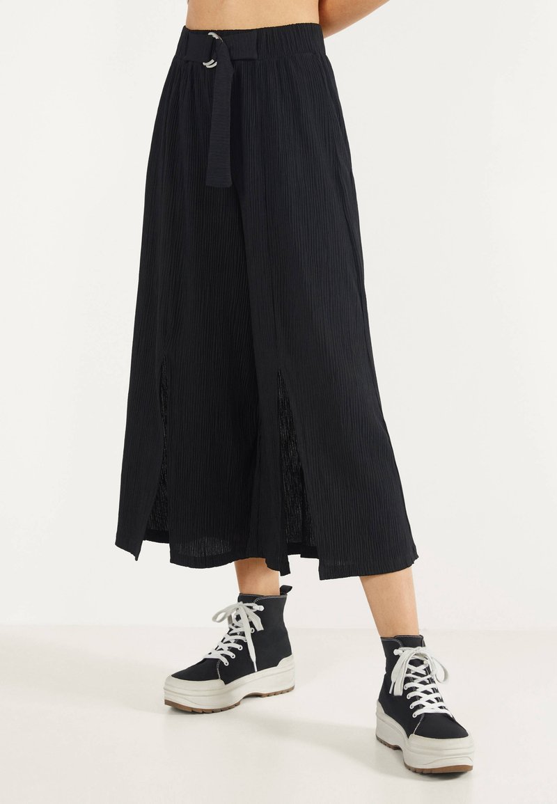 Black pleated maxi skirt with a high waist, a tie belt, and side slits. Paired with black platform sneakers featuring white laces.