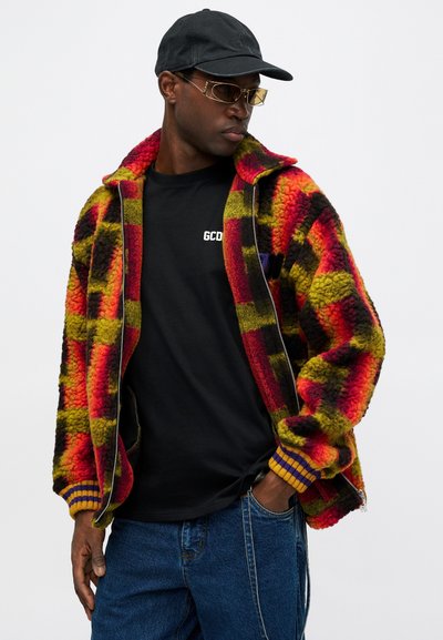 Textūruotas fliso džemperis su raudonai, geltonai ir juodai patikrinamu raštu, su užtrauktuku ir raibais manžetais, dėvimas virš juodos marškinėlių.