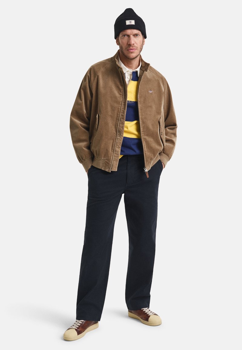 Homem a usar um gorro preto, uma jaqueta castanha, uma camisa riscada azul e amarela, calças azul-marinho e ténis castanhos com bicos em creme. Mãos nos bolsos.