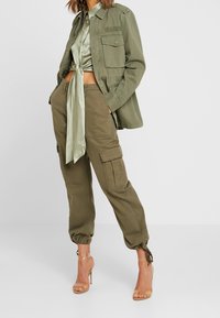 Kvinna som bär olivgröna cargobyxor med ankelband, en knuten satinblus, matchande arbetsjacka och genomskinliga högklackade sandaler.