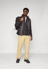 Colete isolante preto por cima de um suéter cinza, calças bege e sapatos escuros. Óculos de sol e uma mochila completam o conjunto.