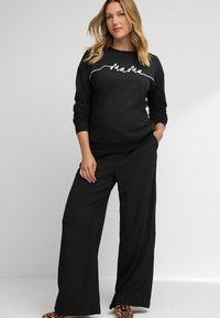 Zwarte sweatshirt met witte tekst "Mama", gecombineerd met zwarte wijduitlopende broeken. Geverfde stof en relaxed fit. Schoenen met dierenprint.