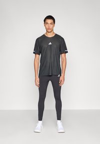 Musta urheilupaita, jossa on tekstuuri ja lyhyet hihat sekä valkoinen logo; yhdistetty mustiin, istuviin leggingsseihin ja valkoisiin lenkkareihin.