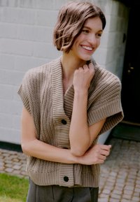 Giovane donna con i capelli corti castano che indossa un cardigan senza maniche di maglia beige, sorridente e in piedi all'esterno vicino a un muro di mattoni bianchi.