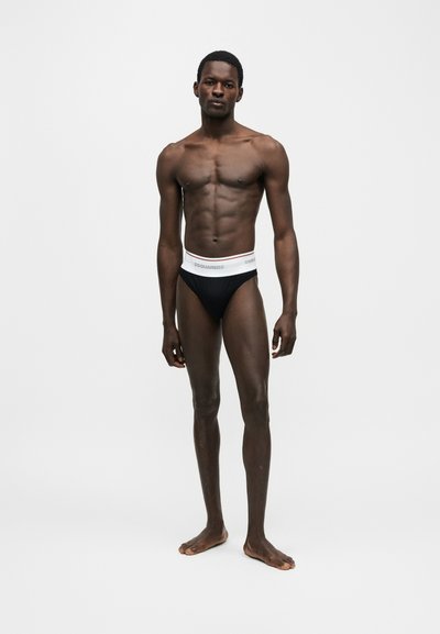 Μυώδης άνδρας στέκεται ξυπόλυτος φορώντας μαύρα σλιπ με λευκή ζώνη που γράφει "DSQUARED2" σε απλό λευκό φόντο.