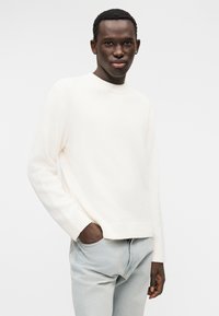 Λευκό, υφασμάτινο πουλόβερ με υφή, με λαιμόκοψη και μανσέτες με ανάγλυφο πλέξιμο, φορεμένο με ανοιχτόχρωμο μπλε τζιν. Τα χέρια του μοντέλου βρίσκονται στις τσέπες, ουδέτερο φόντο.