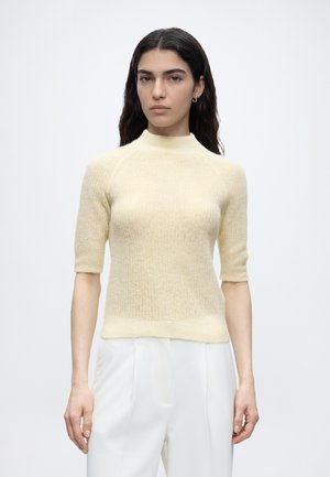 Mujer con cabello largo y oscuro que lleva un suéter de punto corto color crema y pantalones blancos de talle alto sobre un fondo blanco liso.