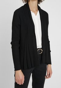 Černý žebrovaný kardigan s otevřeným předním designem, vyrobený z texturované pleteniny a dlouhými rukávy, kombinovaný s bílou košilí a černými kalhotami.