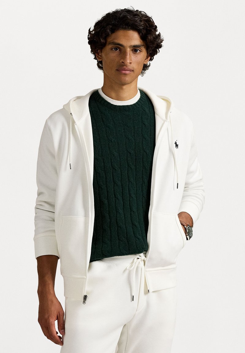 Un hoodie zippé blanc porté sur un pull en maille torsadée vert foncé, avec des poches visibles, et un pantalon blanc ajusté. Le modèle a des cheveux bouclés et porte une montre.