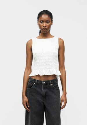 Γυναίκα με πλεξουδωτά μαλλιά, που φοράει λευκό κορυφαίο κοντό τοπ χωρίς μανίκια με βολάν στο τελείωμα και χαλαρό μαύρο τζιν ψηλόμεσο, στέκεται μπροστά σε γκρι φόντο.