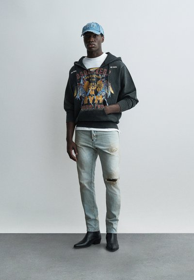 Homme portant une casquette en denim bleu, un sweat à capuche noir avec motif, un t-shirt blanc, un jean clair déchiré et des bottes noires à bout pointu, debout devant un fond uni.