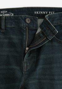 Jeans de denim oscuros de corte skinny con cremallera metálica, costuras contrastantes y etiqueta de marca en la cintura, mostrando un primer plano de la textura de la tela.