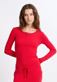 Czerwona długa bluzka henley z rękawami, o prążkowanej fakturze, z okrągłym dekoltem i trzema guzikami na klatce piersiowej.